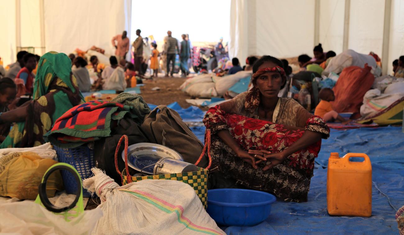 Une femme éthiopienne réfugiée est assise, face caméra, sous une immense tente, sur une grande bâche bleue en plastique. Elle est entourée d’objets divers, dont un couffin, un bidon orange, de la vaisselle, une couverture, une bassine bleue. Derrière elle, en arrière-plan, on aperçoit de nombreux autres réfugiés, hommes femmes et enfants.