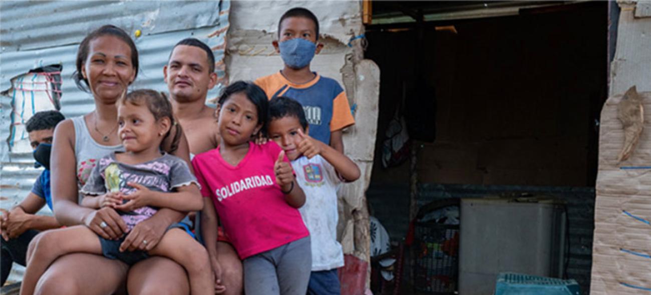 En el exterior de una vivienda humilde, siete miembros de una familia de migrantes venezolanos están todos juntos, posando y miran a la cámara con expresión de agradecimiento. Dos de los cinco menores de edad llevan mascarilla. La imagen fue tomada en la Guajira, Colombia, durante la pandemia de COVID-19.