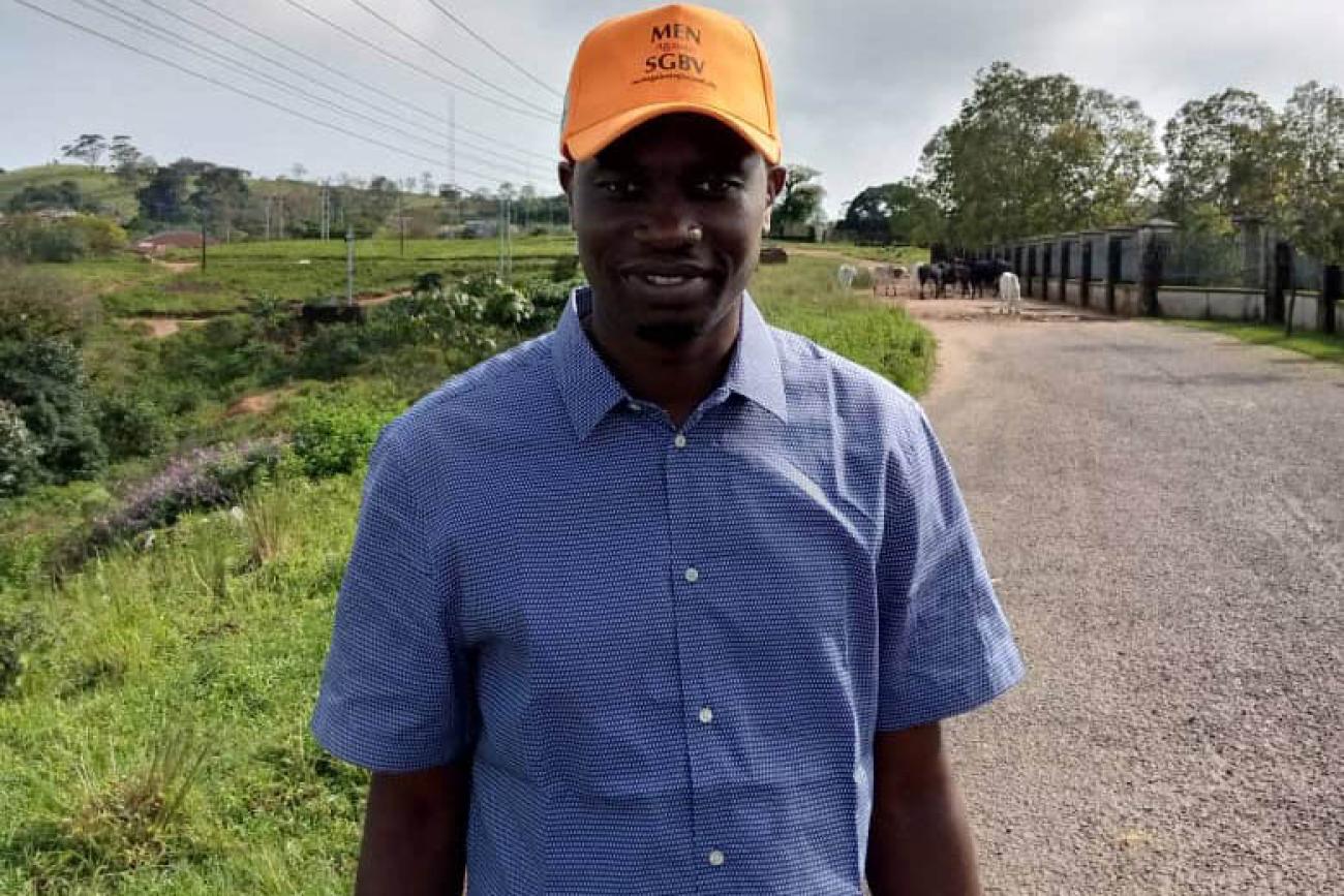 Un hombre, llamando Clement Ejim, sonríe con orgullo a la cámara. Lleva una gorra naranja que muestra su apoyo a la lucha contra la violencia de género. Él está parado en una carretera de cemento cruzando un campo verde.