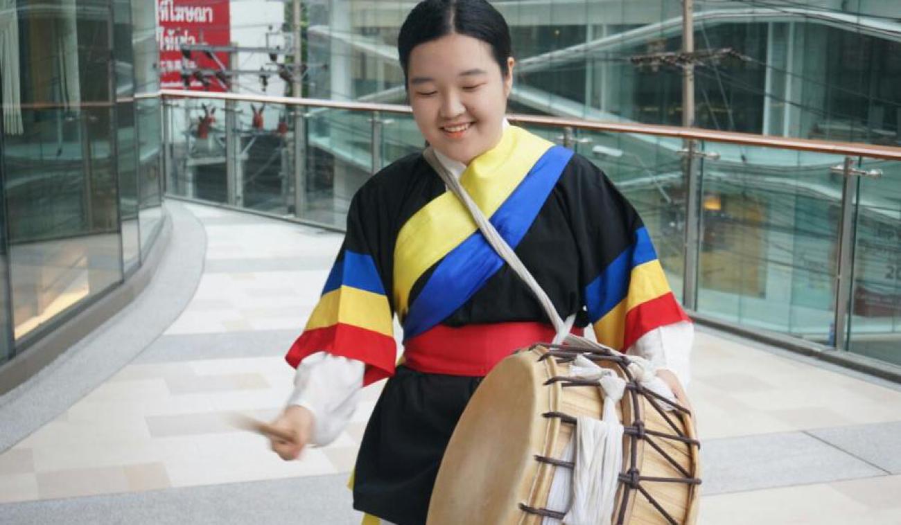 Une jeune fille vêtue d’un habit traditionnel se tient dans les allées d’un grand bâtiment moderne. Elle joue quelques percussions sur un instrument traditionnel, en soutiant.