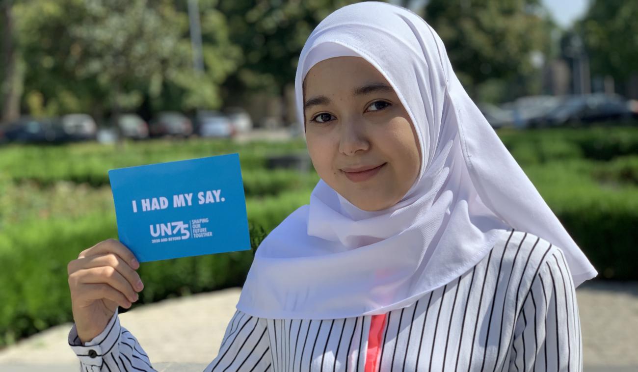 An adolescent girl wearing a head scarf smiles sweetly at the camera as she holds a small blue card that reads "I had my say." 