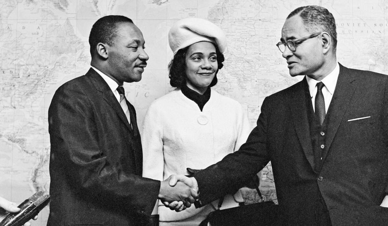 Photo en noir et blanc du Dr Martin Luther King et de son épouse Coretta Scott King accueillis par M. Ralph J. Bunche, Secrétaire général adjoint des Nations Unies aux affaires politiques spéciales, en 1964.