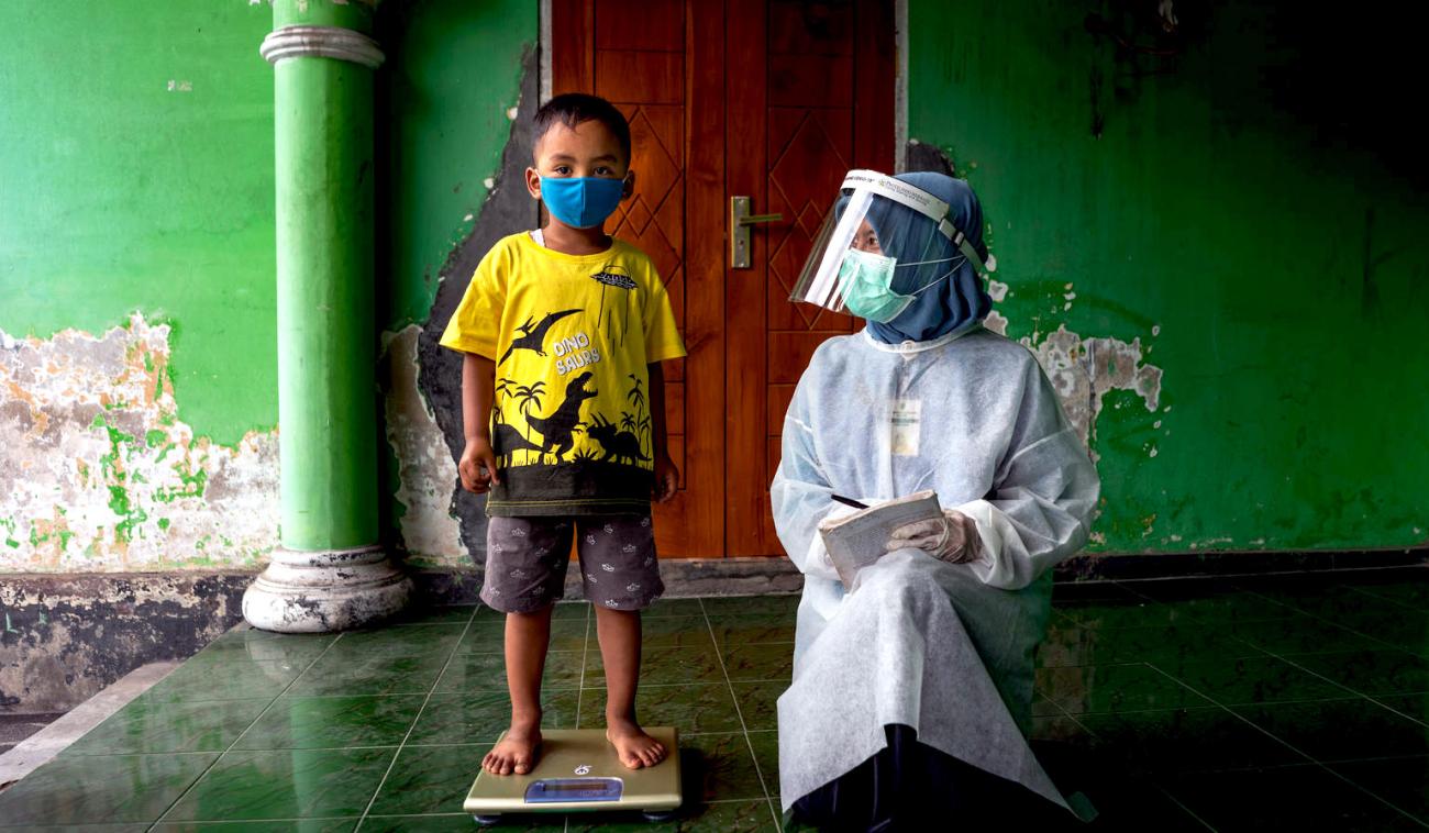 طفل صغير يقف على الميزان وهو ينظر إلى الكاميرا مرتديًا كمامة، ويركع عامل رعاية صحية بجانبه.