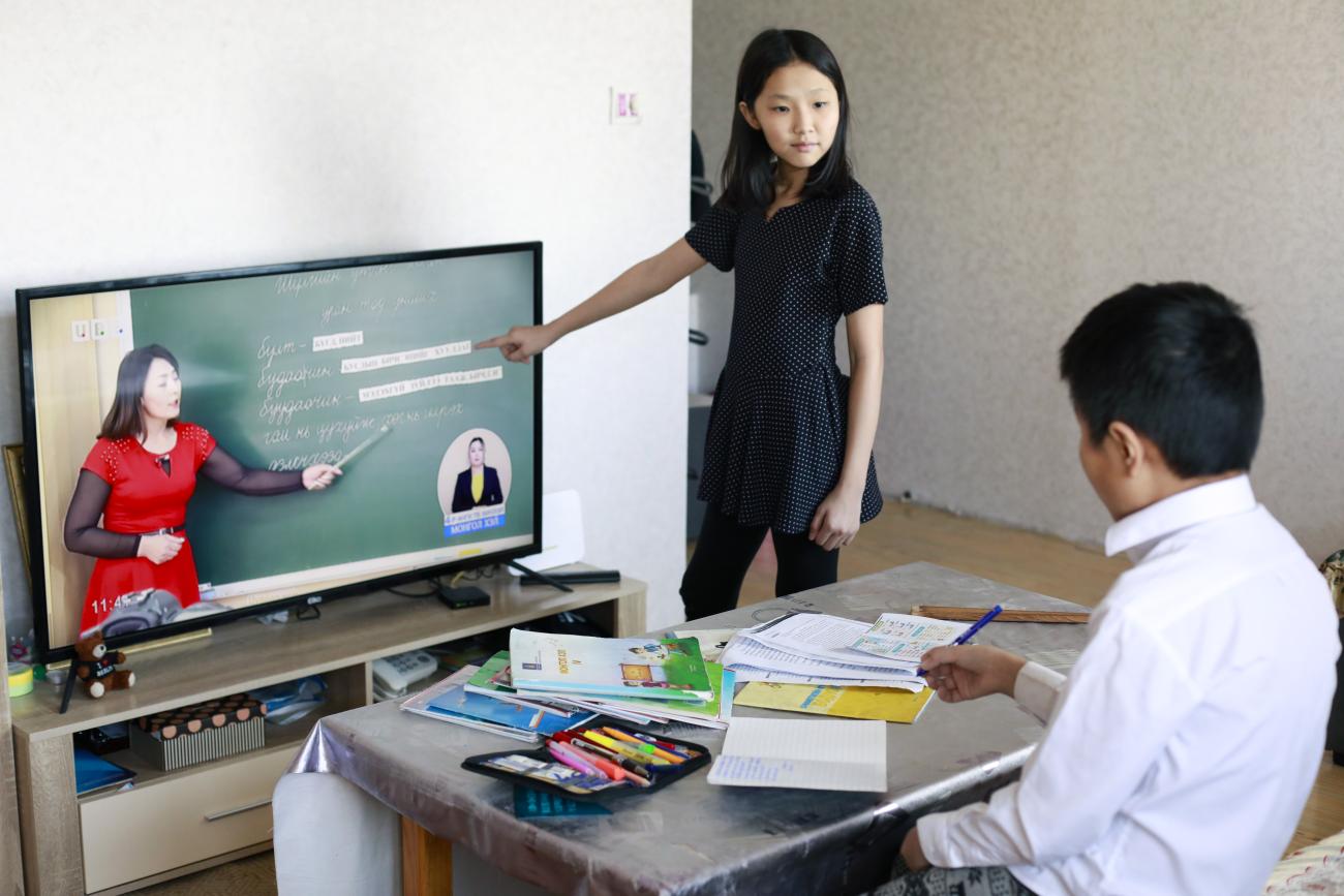 Una niña, con un vestido negro, apunta a una pantalla de televisión que muestra a una maestra, mientras que un niño con una camisa blanca toma notas junto a una caja de lápices.