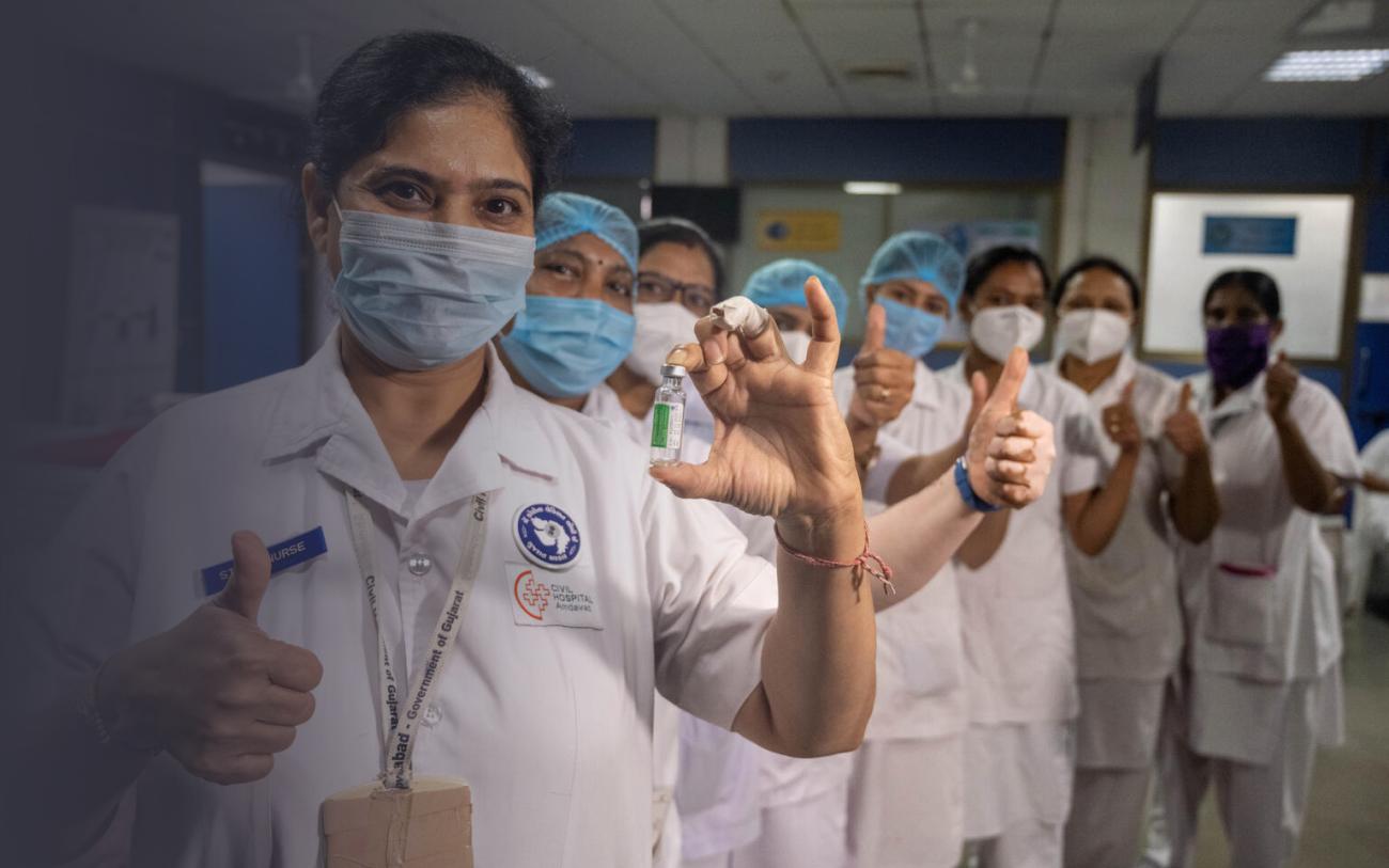 Работники больницы, стоя в ряд в больничном коридоре, показывают поднятый вверх большой палец в знак одобрения, женщина на переднем плане показывает в камеру ампулу с вакциной.