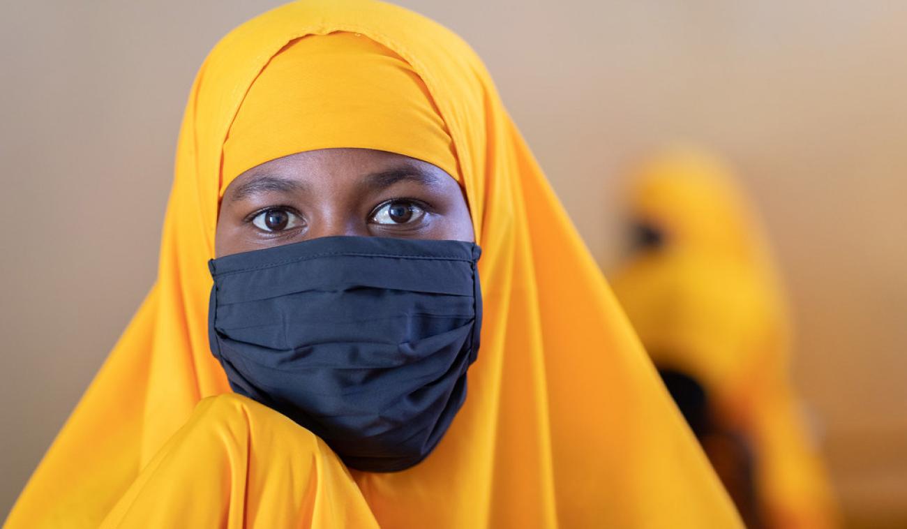 A young adolescent girl wearing a yellow headdress and black mask looks strikingly at the camera.