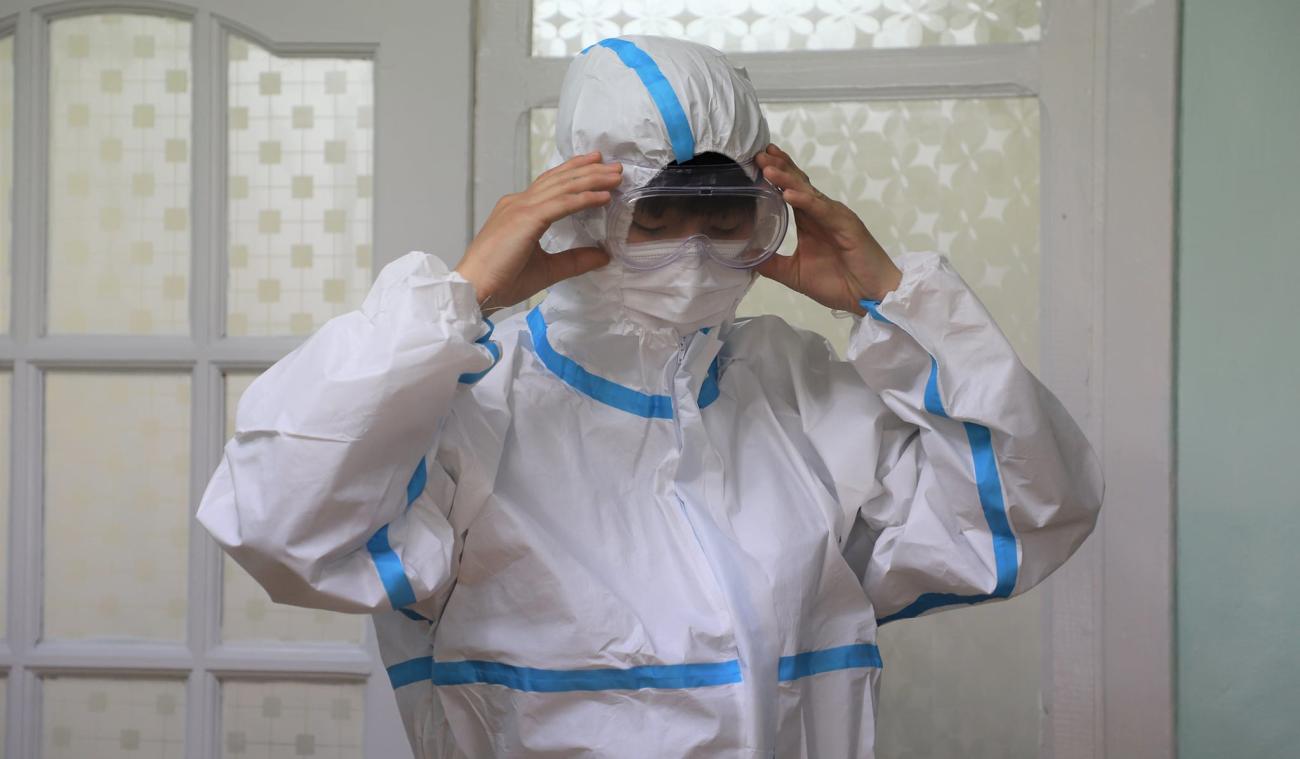 Un membre du personnel de santé portant un équipement de protection individuelle de la tête aux pieds ajuste son masque de protection en plastique sur le visage.