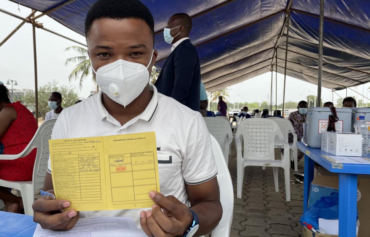 A man in a white shirt and white face mask holds up a yellow COVID-19 vaccination card. 