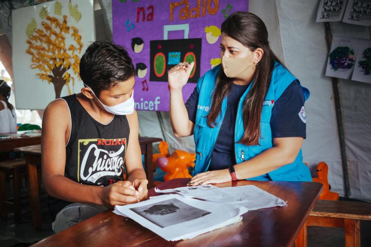 A teacher in a blue vest and face mask talks to a boy in a black tank top and white face mask. 