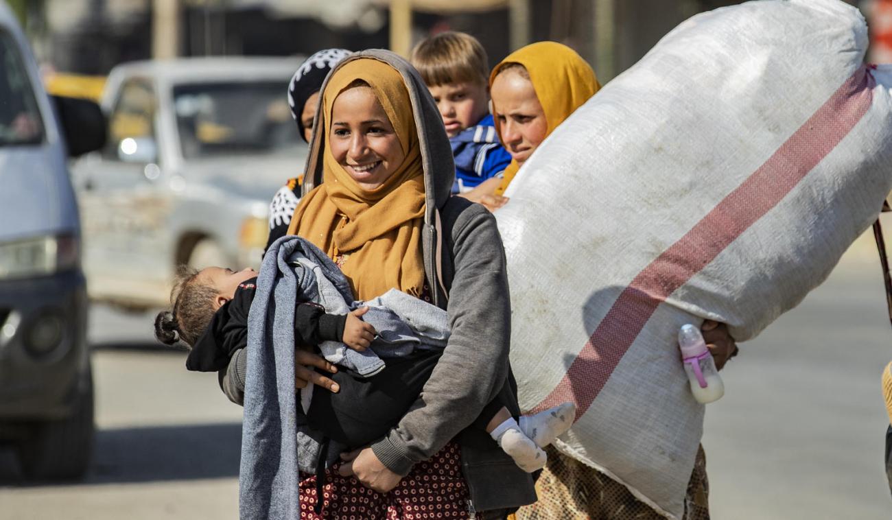 A woman smiles as she carries a sleeping child. Another woman walks behind her as she carries a large bag. 