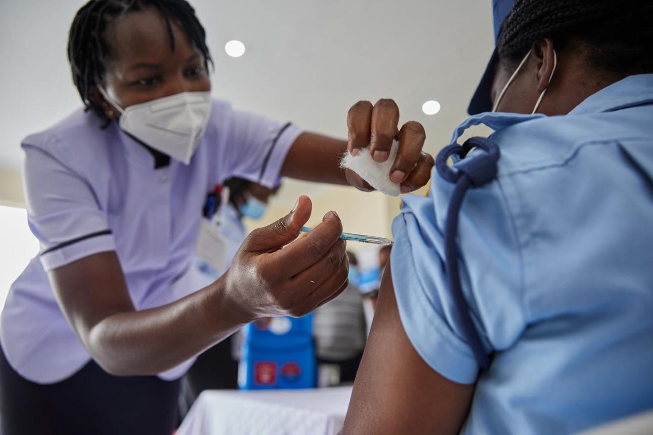 Una enfermera con una camisa morada y una mascarilla blanca administra una vacuna contra el COVID a una mujer con una camisa azul y gorra.