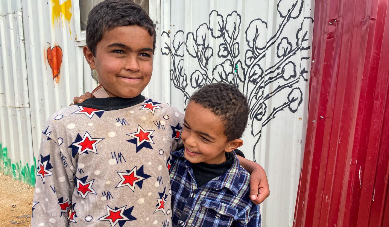 Dos niños pequeños abrazados sonríen felices mientras están parados afuera, uno al lado del otro, junto a una pared de zinc que tiene una pintura mural, con la silueta de un árbol.