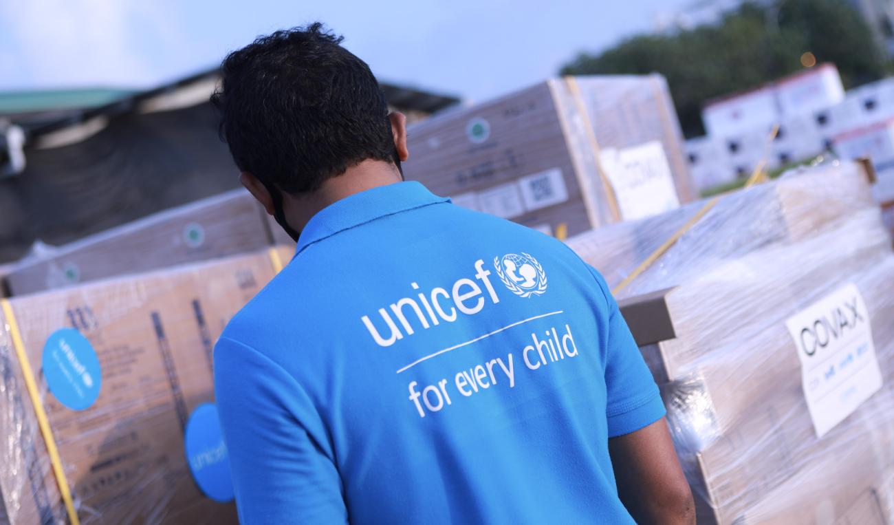 Un hombre con una camiseta azul claro de UNICEF observa la entrega de vacunas envueltas en papel de regalo.