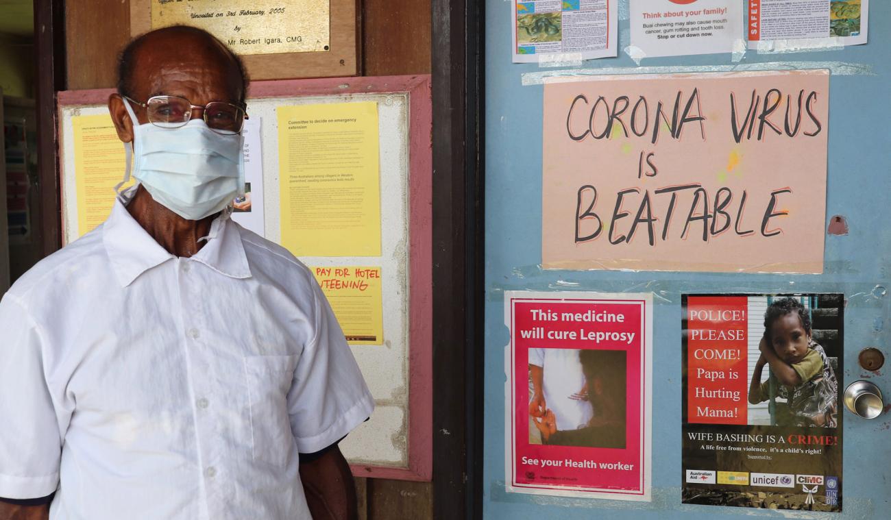 Un homme vêtu d'une chemise blanche et portant masque de protection respiratoire blanc se tient près d'un panneau d'affichage où figurent des informations sur la COVID-19 et une inscription indiquant "Corona virus is beatable", ce qui signifie en français "Nous pouvons vaincre le coronavirus".