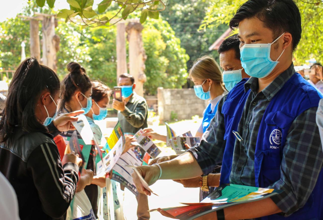 Plusieurs membres du personnel de l'OIM portant des vestes bleues et des masques de protection distribuent des brochures d'information à un groupe de jeunes femmes qui portent elles aussi des masques de protection.