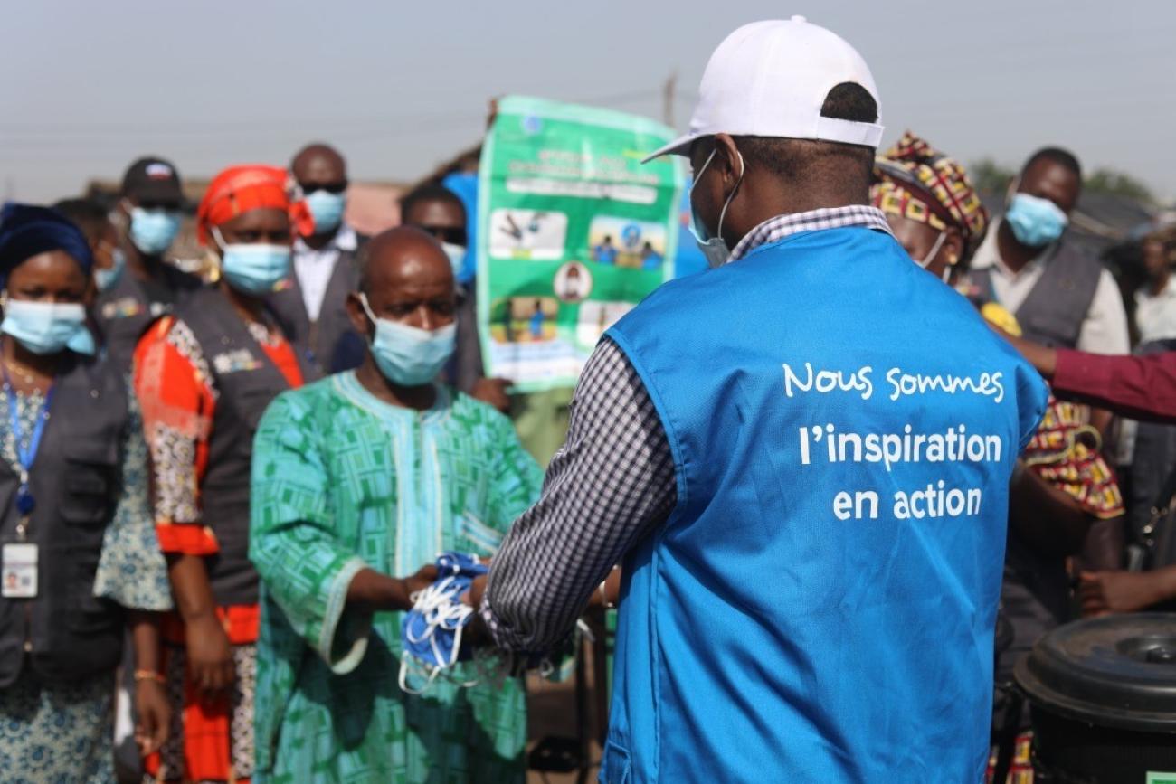 A United Nations Volunteer in a blue vest hands a man a bag. Several people stand behind the man receiving the package.