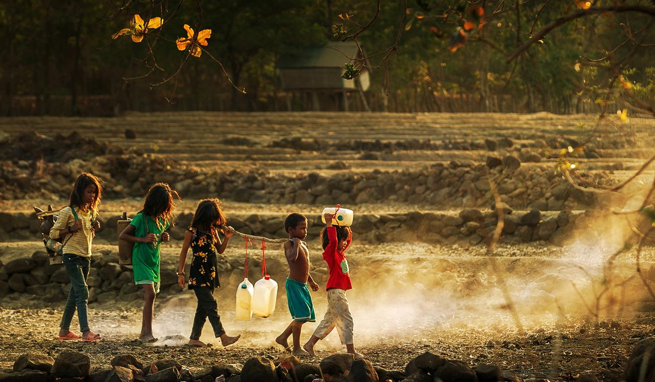 Cinco niños y niñas caminan en fila sobre un terreno árido, y entre todos transportan garrafas con poco líquido adentro.