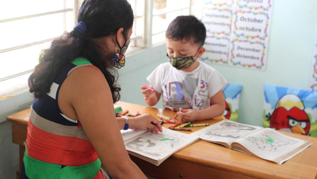 A woman and a child in face masks look over coloring books on a wooden table. 