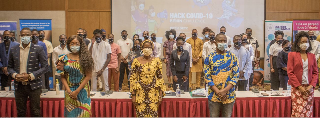 A large group of people in face masks stand facing the camera in front of projector displaying the words "Hack COVID-19."