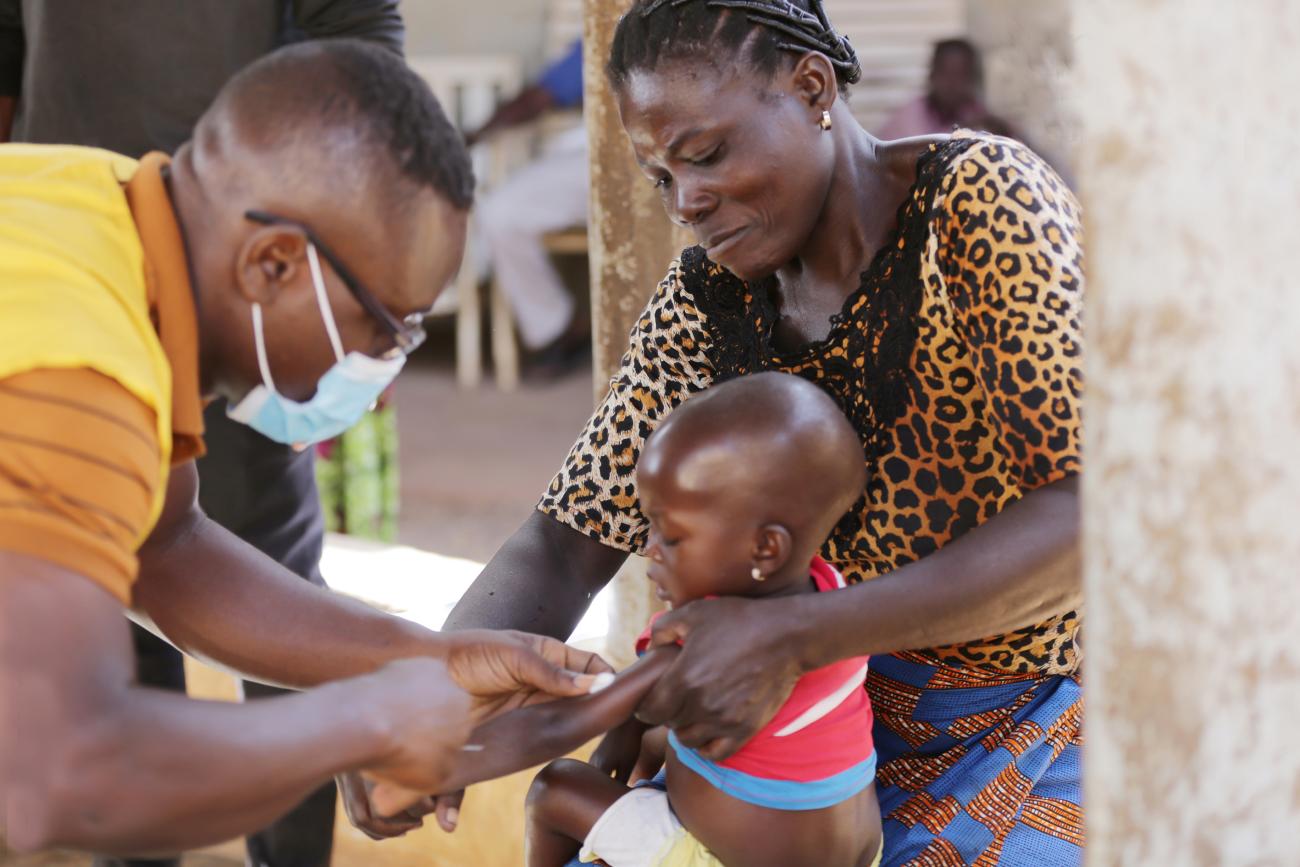 A child, held by a woman, receives an injection from a man in a yellow shirt. 
