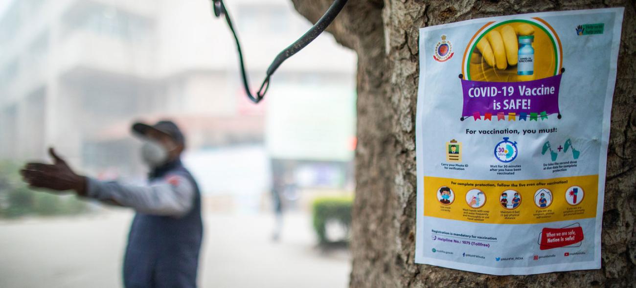 A sign, with the image of a vaccine and information, is posted on a tree and a person in a mask is seen in the background pointing. 