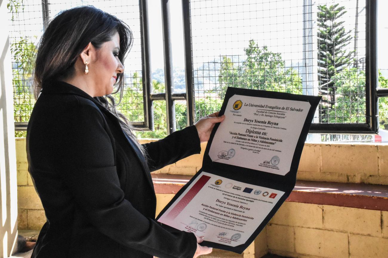 A woman with dark hair and a black jacket looks down at her diploma.