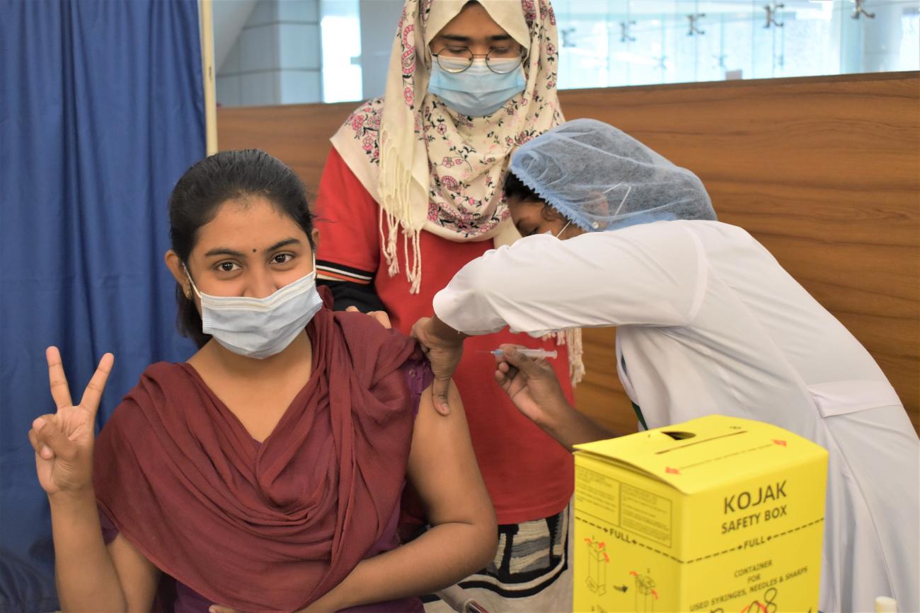 Une jeune femme se fait vacciner contre la COVID-19 en faisant avec les doigts le signe « V », pour « Vaccin ». Une femme portant une blouse blanche et une charlotte bleue lui administre le vaccin. Une troisième femme portant un foulard se tient debout en arrière-plan. Les trois femmes portent un masque de protection.