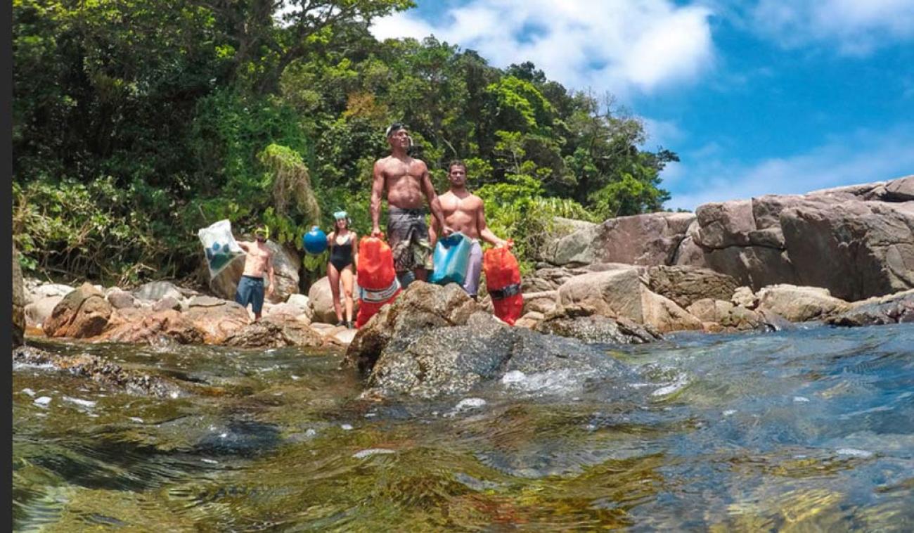 Cuatro personas, en traje de baño y con equipamiento para realizar una limpieza, se paran sobre rocas cerca del océano.