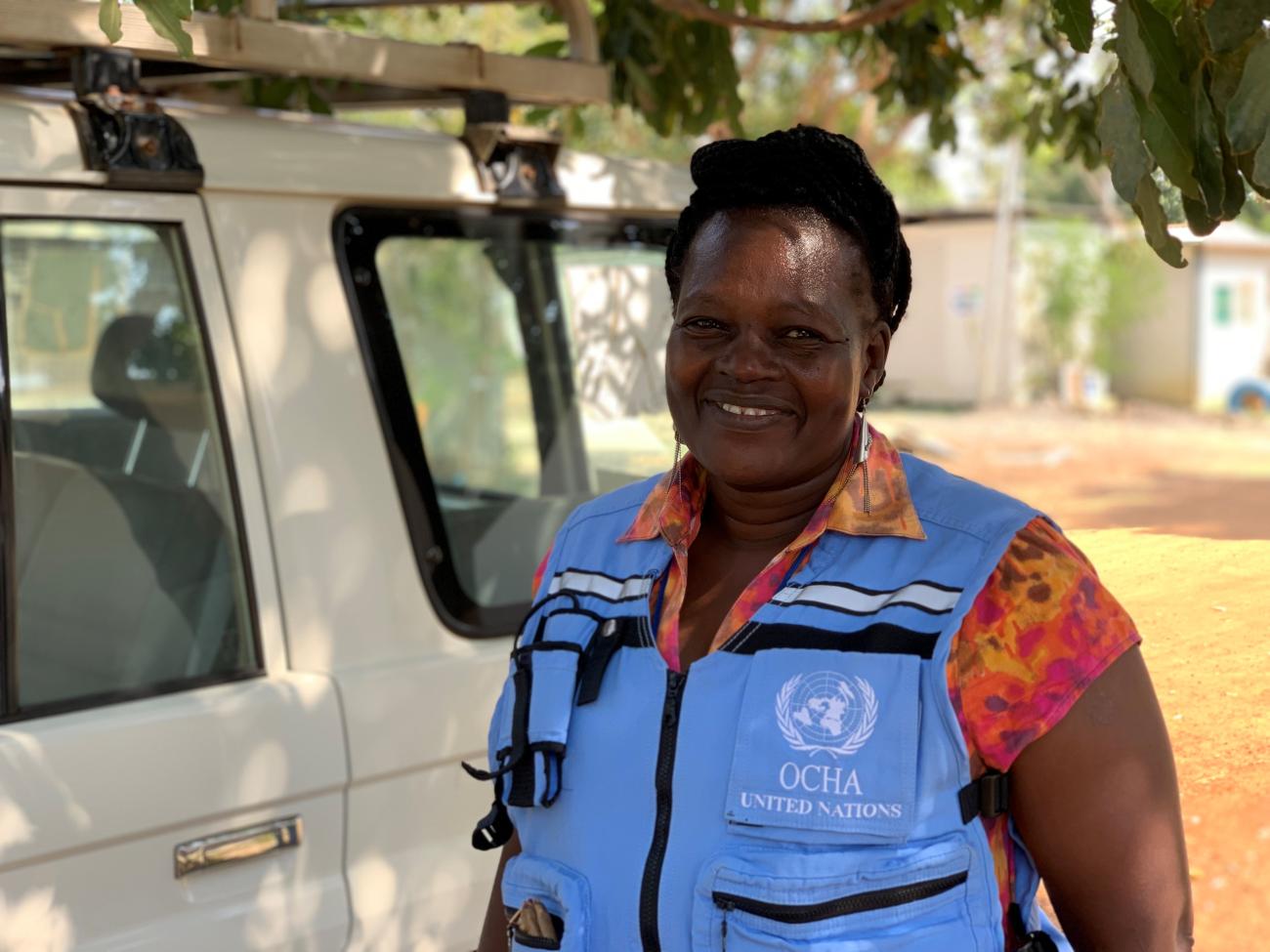 Une femme portant un gilet bleu clair de l'OCHA, le Bureau des Nations Unies pour la coordination des affaires humanitaire, sourit à la caméra.