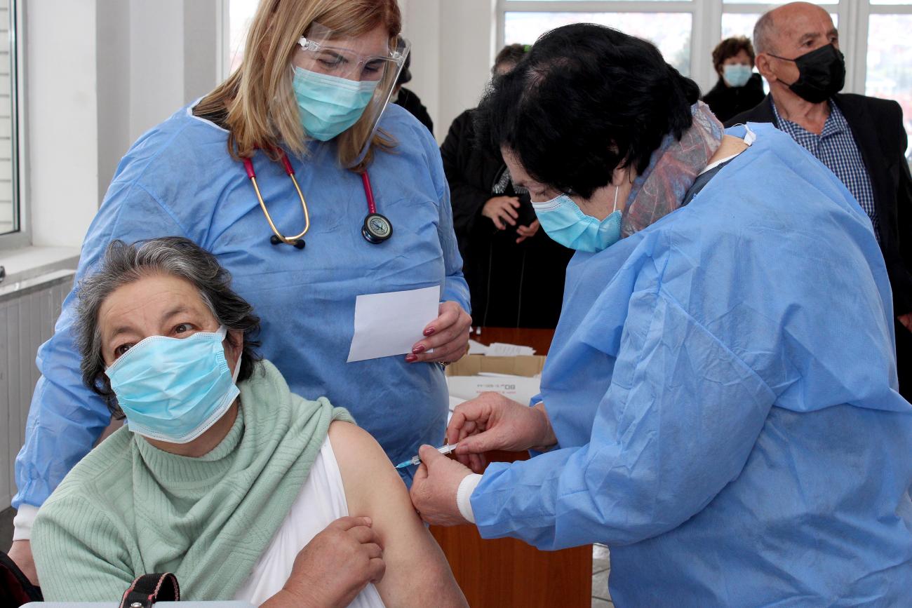 Une femme portant un masque de protection est assise et tend le bras gauche pour se faire vacciner. Deux femmes portant une blouse bleue et un masque se tiennent debout, penchées sur elle. L’une d’elle lui administre le vaccin tandis que l’autre la regarde réaliser le geste.