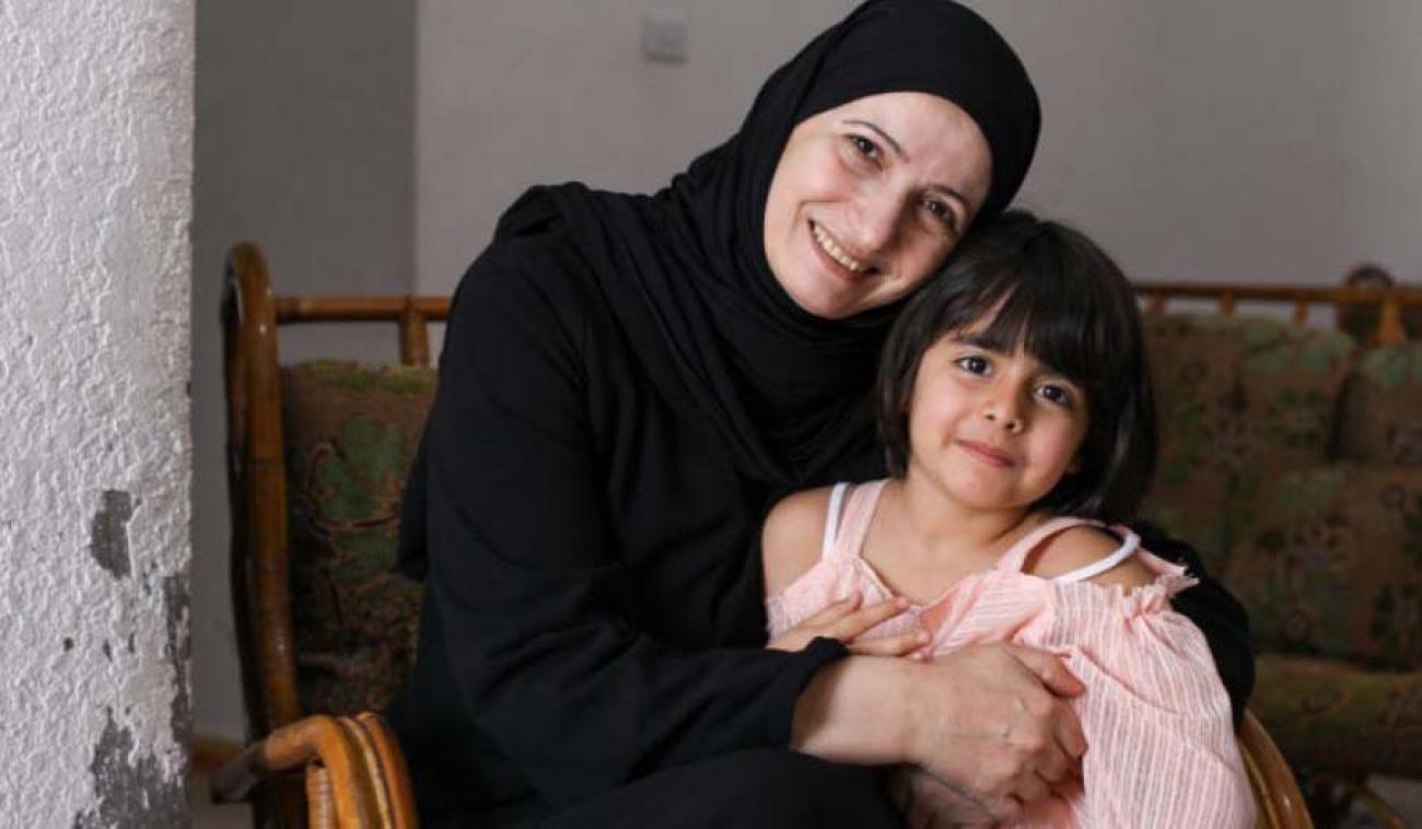 Ghada smiles cheerfully as the camera as she proudly embraces her youngest daughter, who is also smiling at the camera.