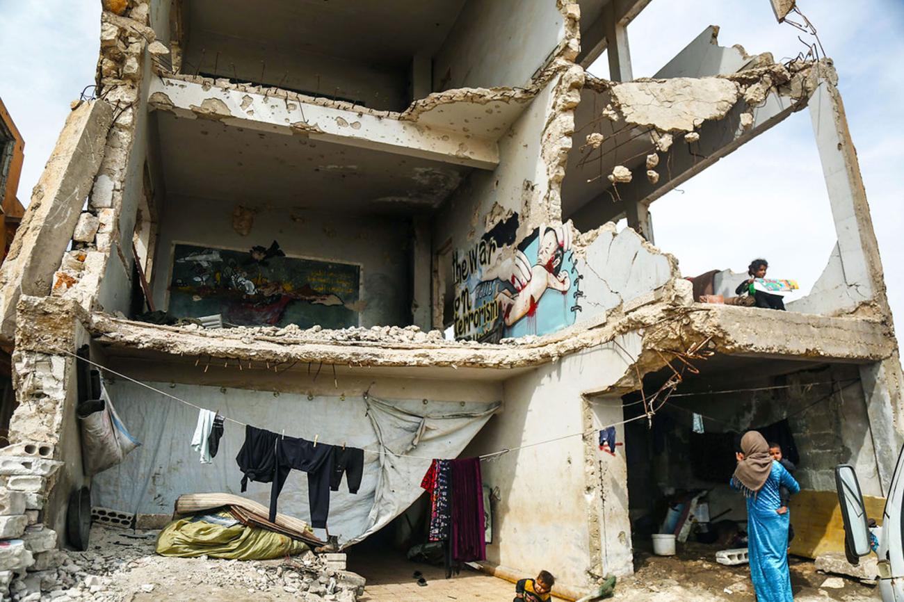امرأة في ثوب أزرق وأطفال يقفون داخل وقرب مبنى مدمر جزئيًا.