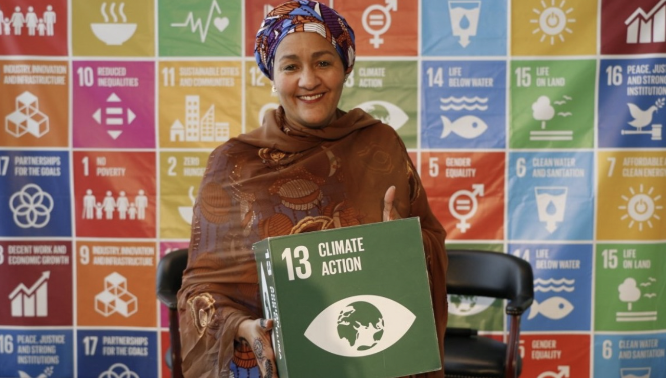 Amina Mohammed, Vicesecretaria General de la ONU, sonríe a la cámara mientras sostiene una caja verde con las palabras "Climate Action", en inglés (las cuales se traducen como "Acción por el Clima", en español).