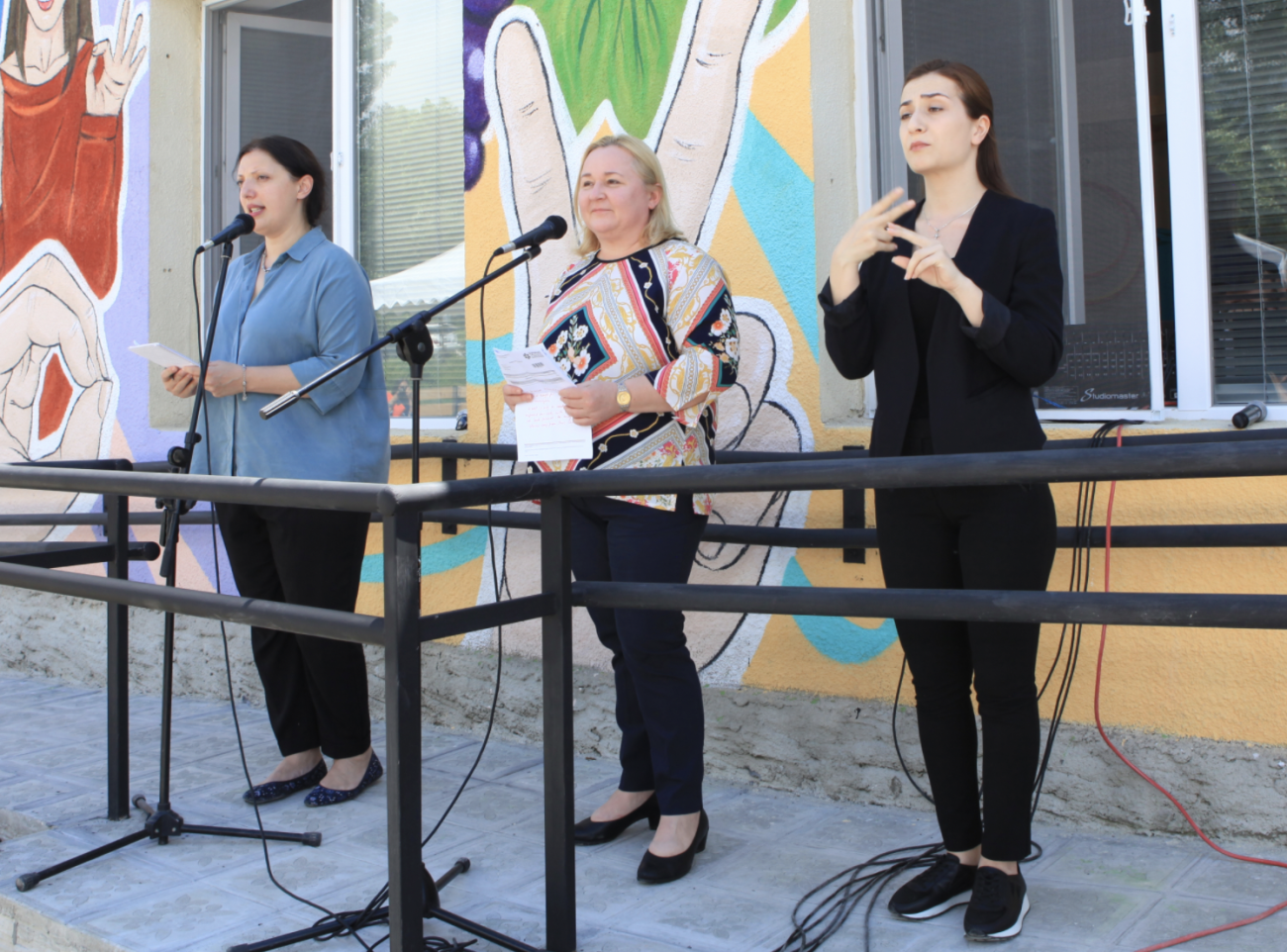 Trois femmes se tiennent debout devant un centre d'inclusion sociale dont la façade est recouverte de peintures murales colorées. Une femme, à l'extrémité gauche de l'image, s'exprime au micro, tandis qu'une autre femme, à droite de l'image, traduit le discours prononcé dans la langue des signes.