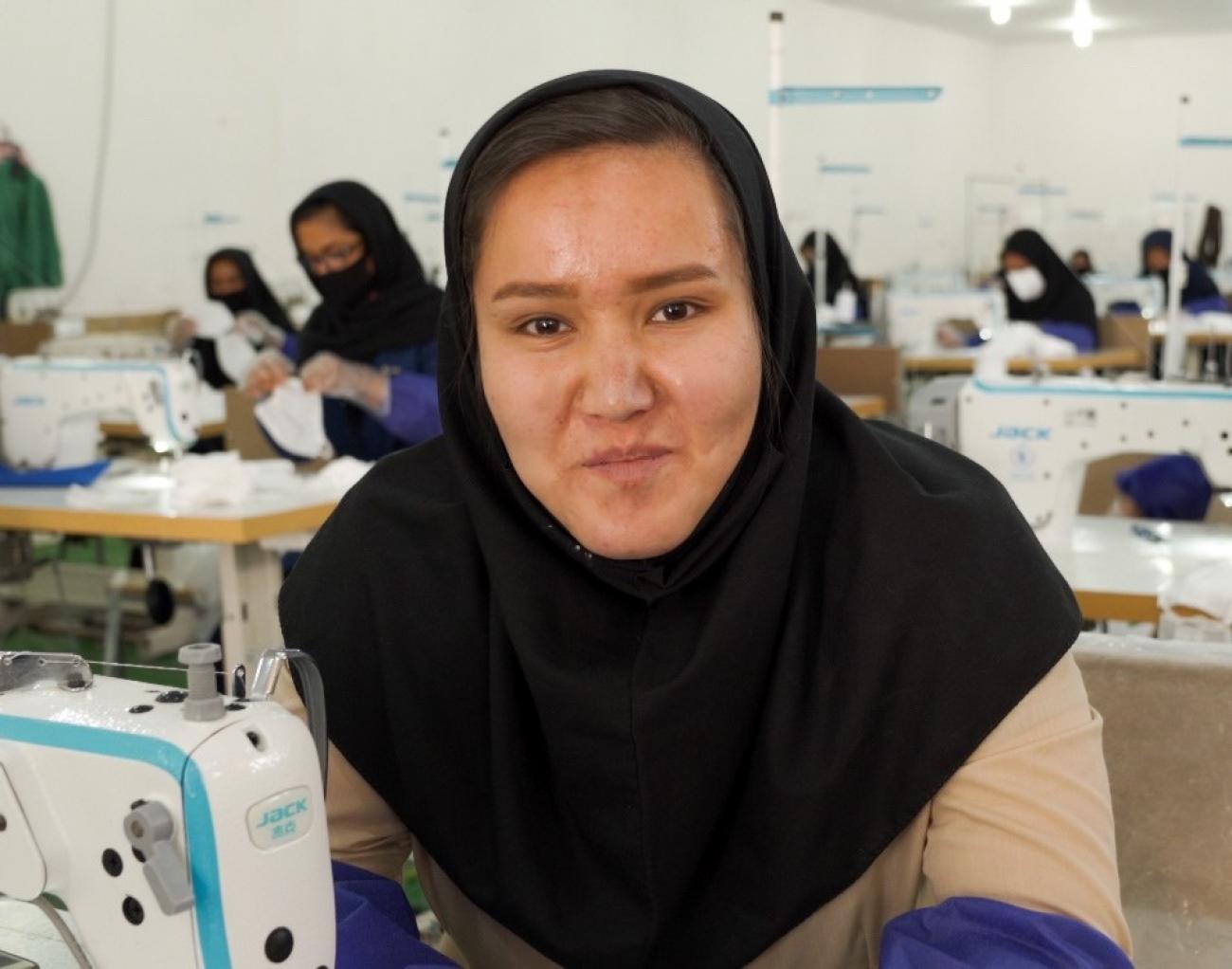 A young woman looks directly at the camera in a room full of women working at sewing machines. 
