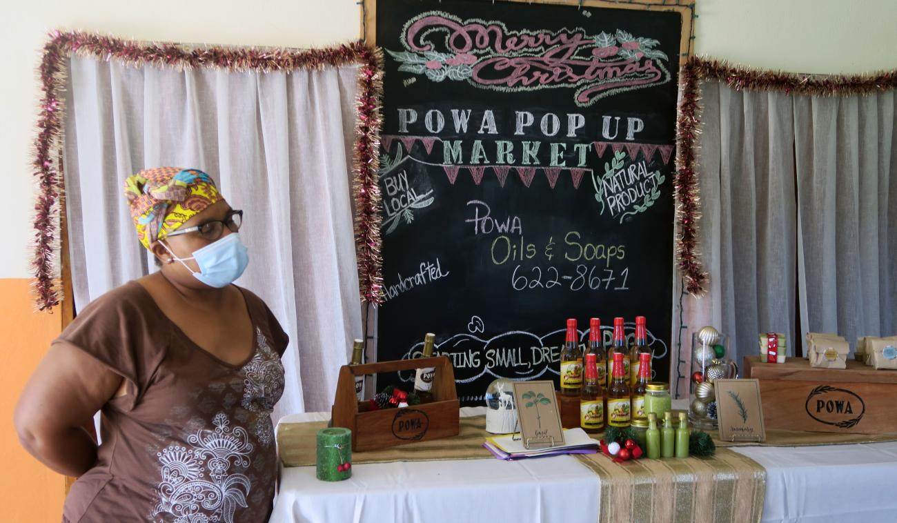 A woman wearing a face mask stands in front of her homemade oils and soaps at a POWA-sponsored market.