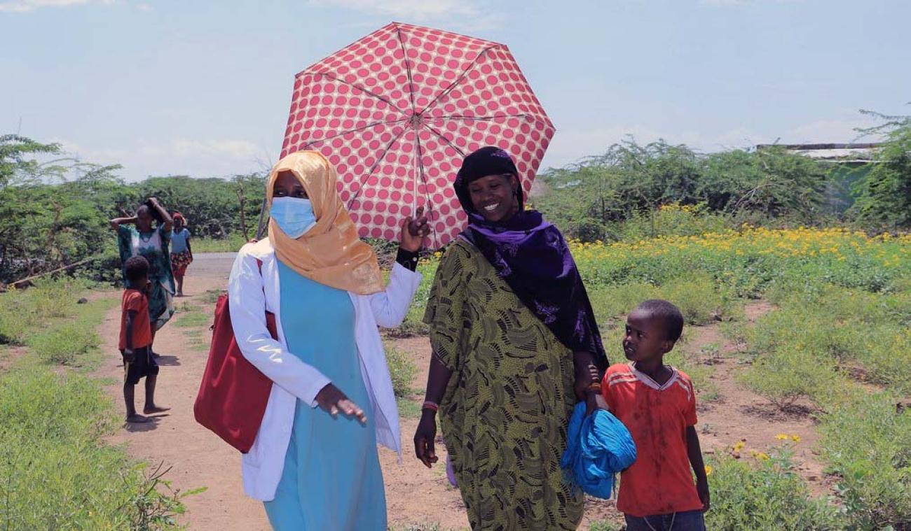 One woman wearing a face masks holds an umbrella as she walks side-by-side with a woman and her child.