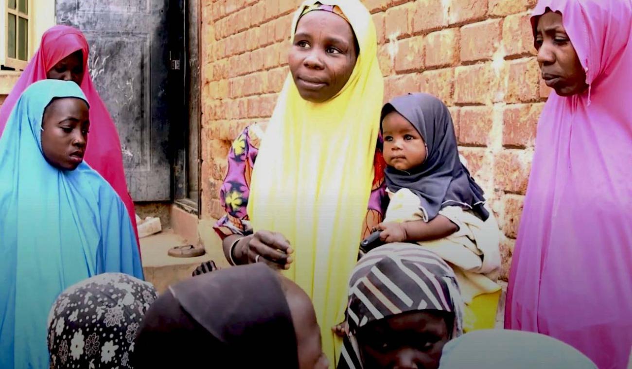 Una mujer con un hiyab amarillo sostiene a un bebé en brazos mientras habla con otras mujeres.
