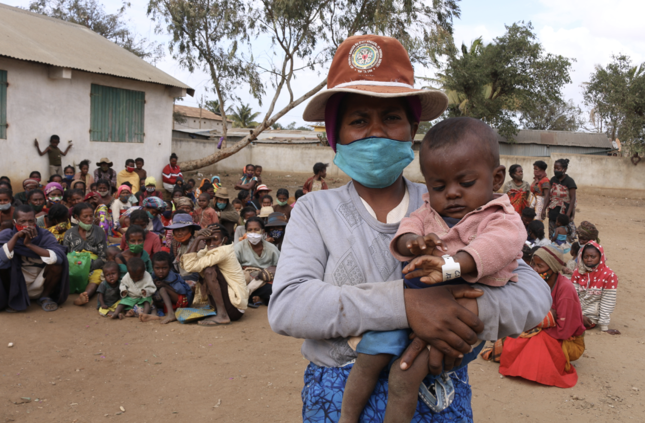 一个戴着面具和帽子的妇女怀里抱着一个婴儿，一大群人坐在她身后。