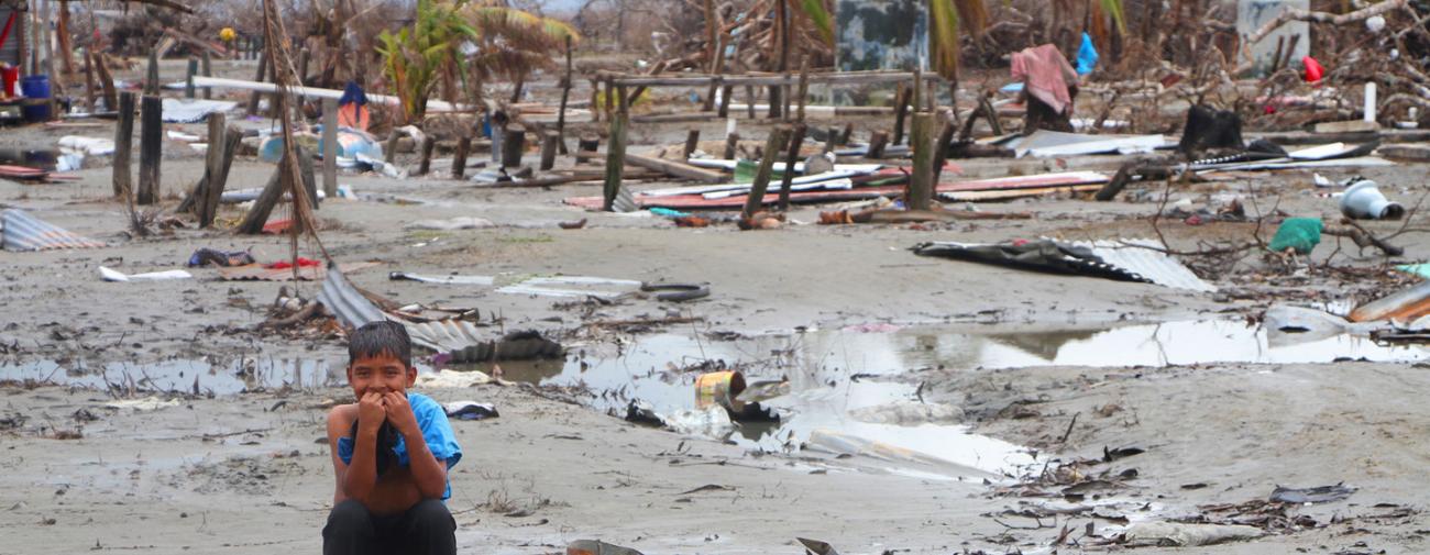 Un niño en primer plano, con un fondo de barro a sus espaldas, entre los escombros de lo que fue su hogar.