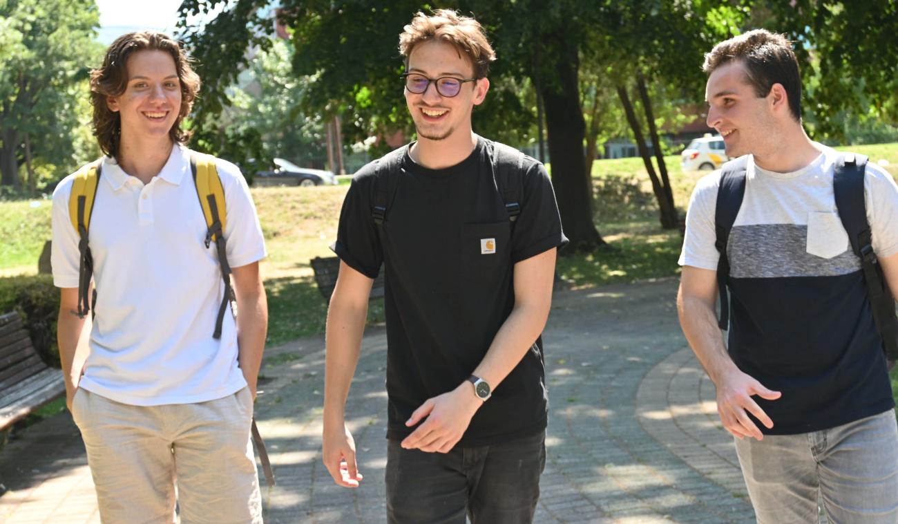 Trois jeunes hommes, Teo (à gauche), Orhan (au milieu) et Boris (à droite), marchent côte à côte en souriant dans un parc.