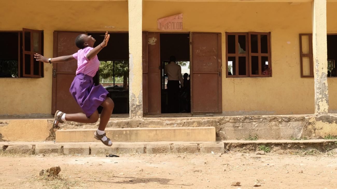 A young girl in a pink shirt and purple dress jumps near a school building.