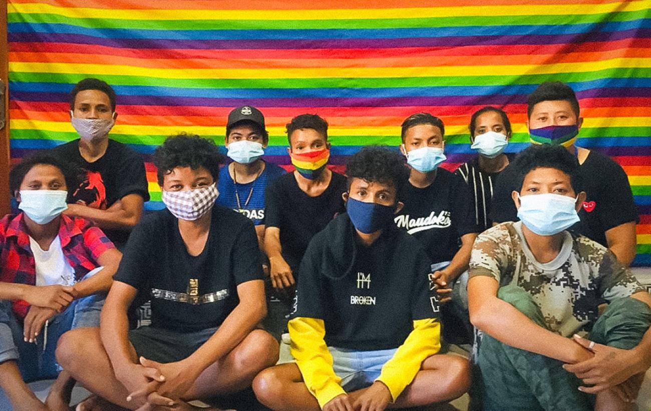 A group of young people wearing masks sit cross-legged in front of a rainbow-coloured flag.