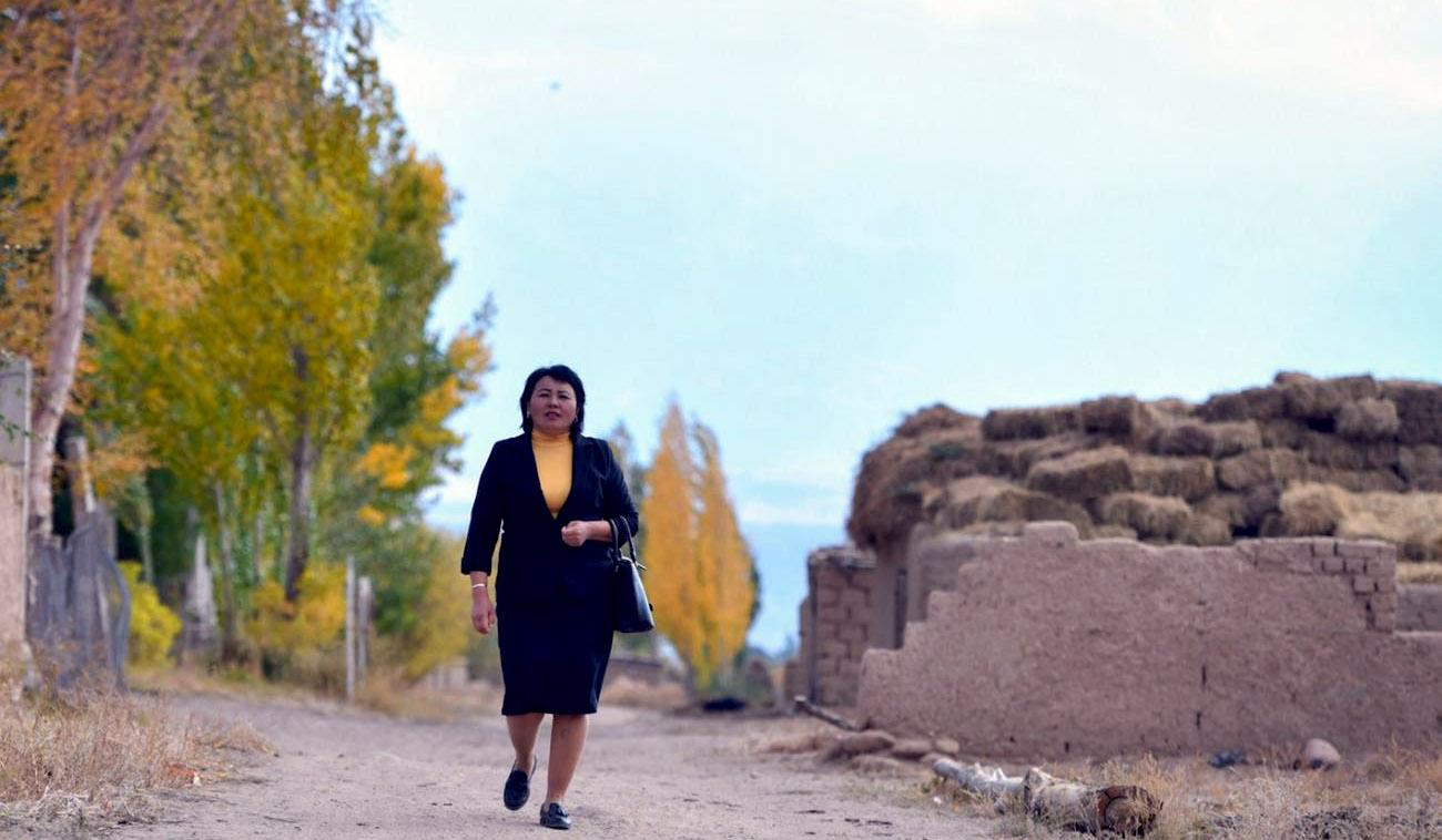 A woman in a black suit and yellow shirt walks proudly down a road towards the direction of the camera.
