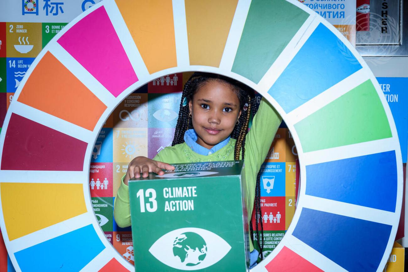A young girl, Eva Mendes de Leon, smiles at the camera as she poses in the centre of a large SDG wheel as she holds a blog with the SDG13 logo.