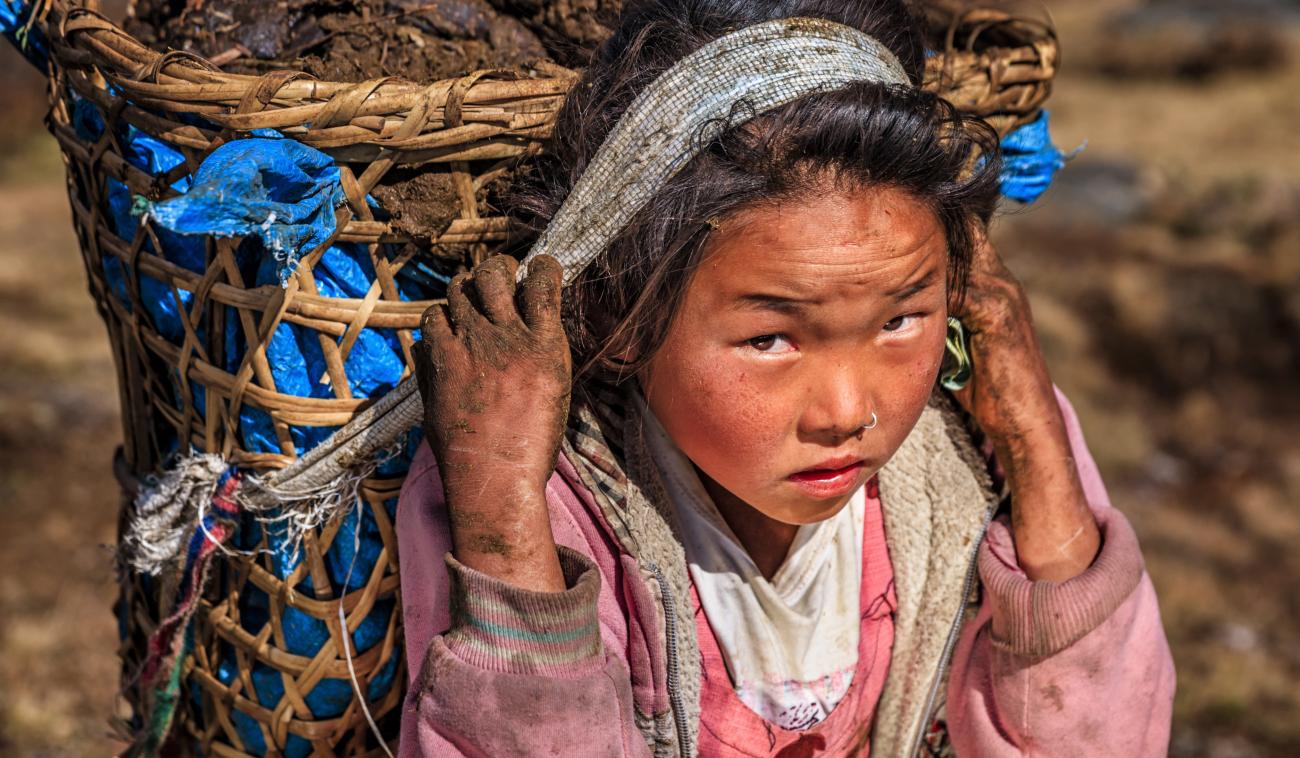 Una joven, con sucio en sus manos y su rostro, lleva una gran cesta a la espalda.