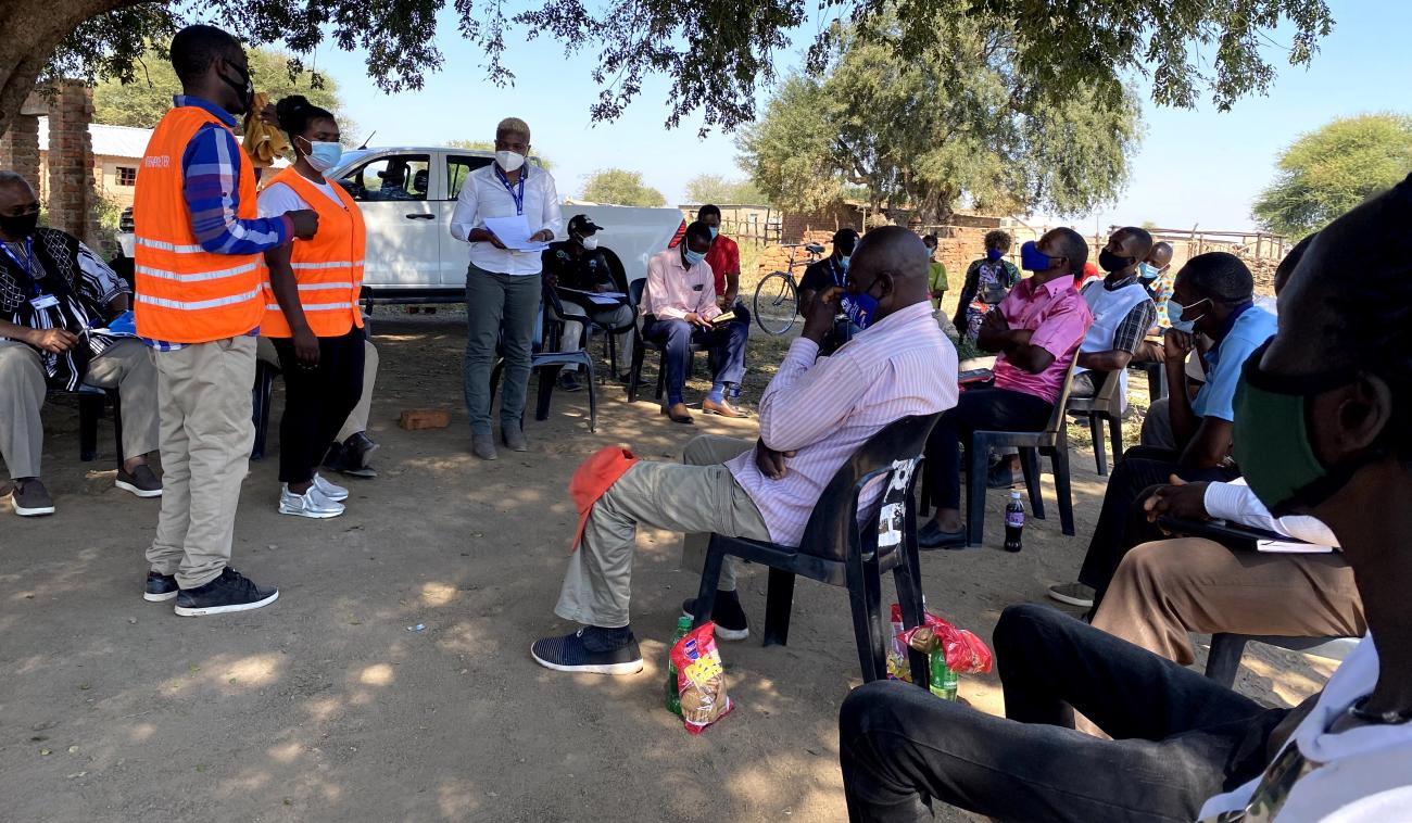 Des membres du personnel de l'ONU se tiennent debout devant un groupe de réfugiés et de demandeurs d'asile assis en groupe, sur des chaises en plastique, à l'ombre d'un arbre, dans le camp de Tongogara. Toutes et tous portent un masque de protection respiratoire.