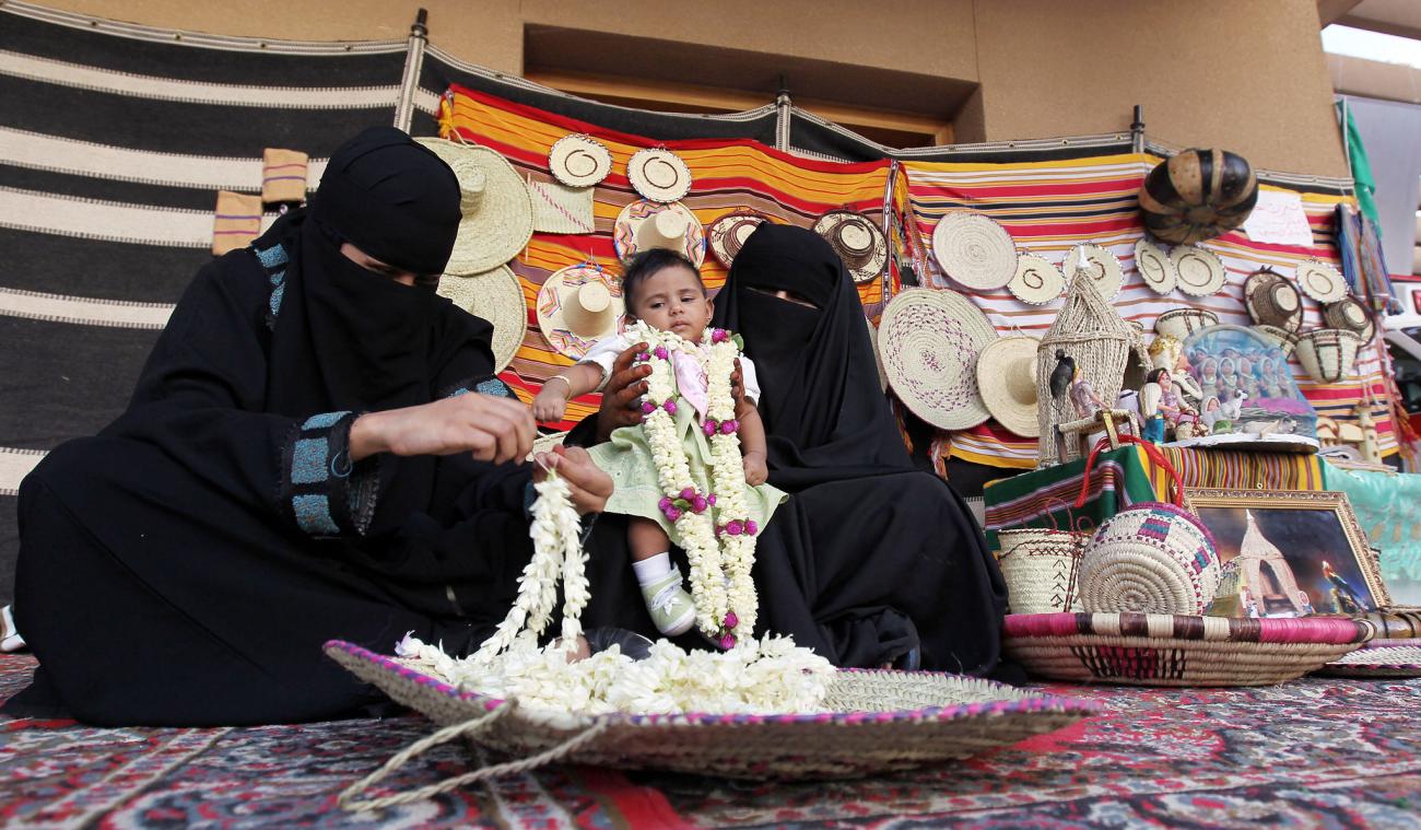 A Saudi woman sits on a rug and threads fresh flowers to sell in a souk as another woman sitting beside her holds up a baby. 