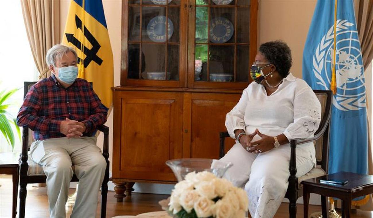 La Première ministre de la Barbade (à droite) et le Secrétaire général des Nations Unies (à gauche) sont assis l'un à côté de l'autre sur des chaises en bois, devant les drapeaux de l'ONU et de la Barbade. Ils portent tous deux un masque de protection respiratoire.