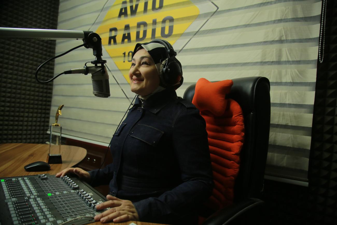 Une femme vêtue d'une tenue bleu foncé et portant un foulard sur la tête et un casque aux oreilles est assise à un bureau, dans un studio radio, devant un microphone et regarde devant elle en souriant.